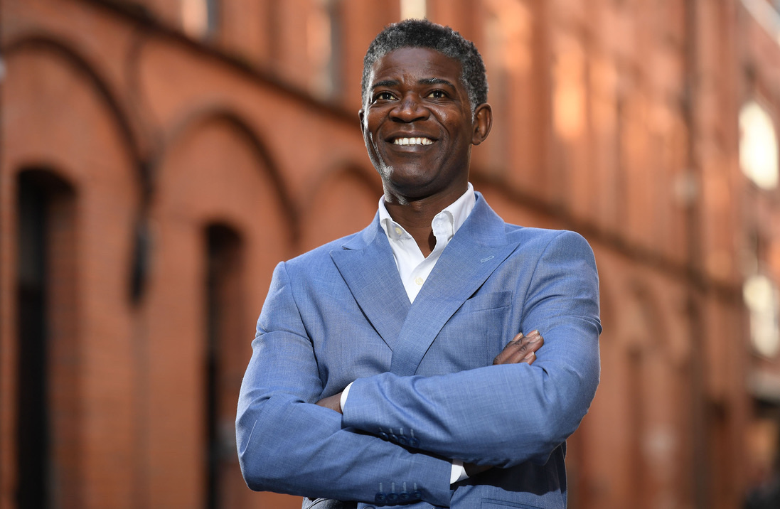 Tura Arutura in a blue suit, arms folded and smiling, on a red-brick Belfast street