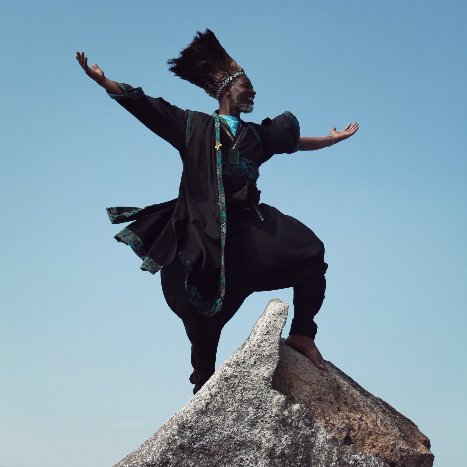 Tura Arutura in ceremonial dress with a feathered headdress, arms outstretched on a granite rock against a blue sky